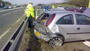 a_police_traffic_officer_in_a_hi-viz_jacket_standing_by_a_central_reservaton_motorway_barrier_inspecting_a_damaged_car_which_has_collided_with_the_barrier